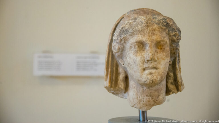 Weathered marble female head with center-parted hair and a veil or mantle descending along both sides of the face.