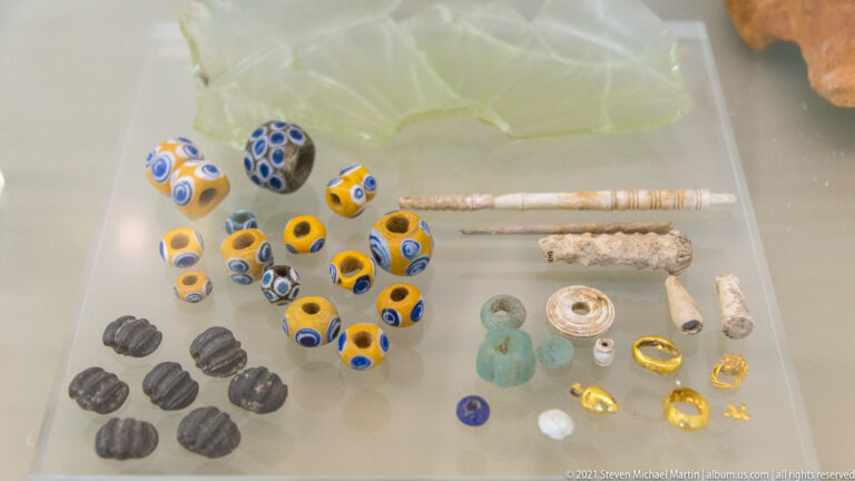 Museum case with yellow-blue glass beads, dark ribbed beads, green glass pieces, slender pins, and small gold rings and fittings.