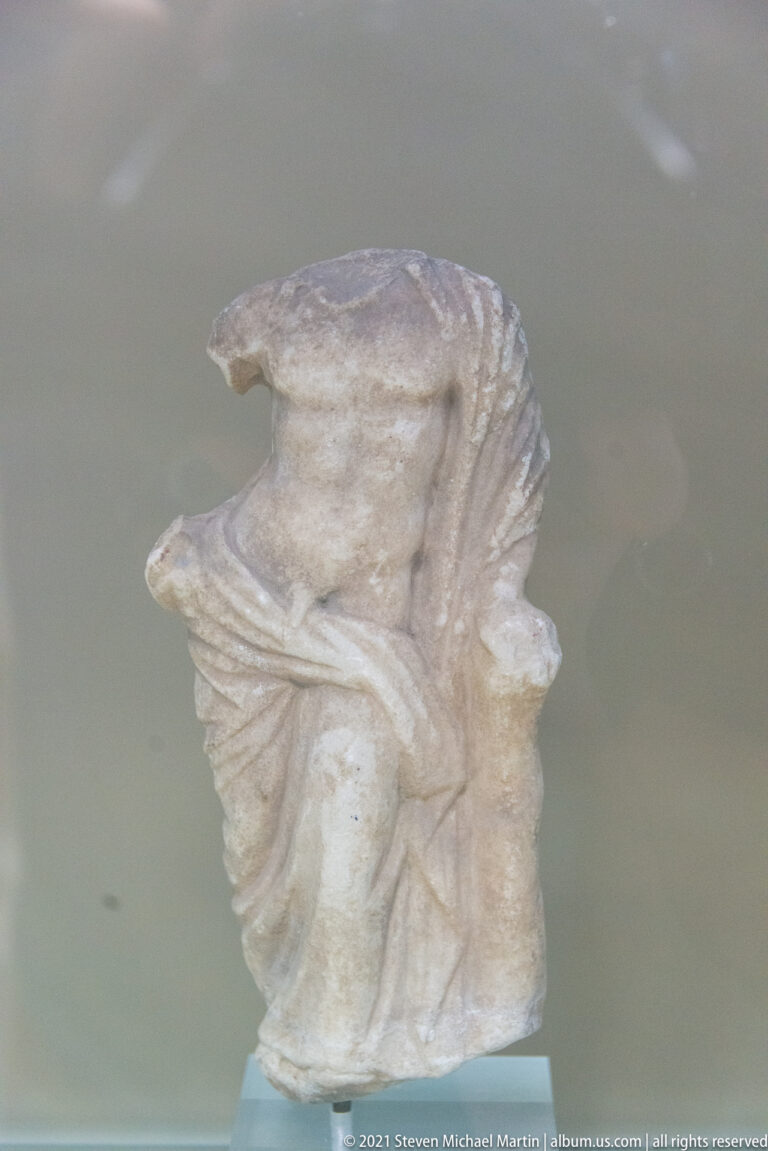Headless and limbless marble torso fragment with deep diagonal folds of drapery wrapping the upper body.