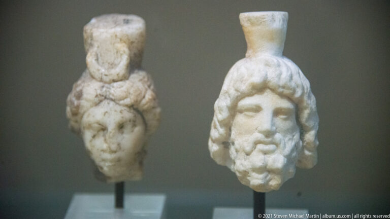 Two small pale stone heads on mounts, the front one a bearded long-haired male and the rear one more fragmentary.