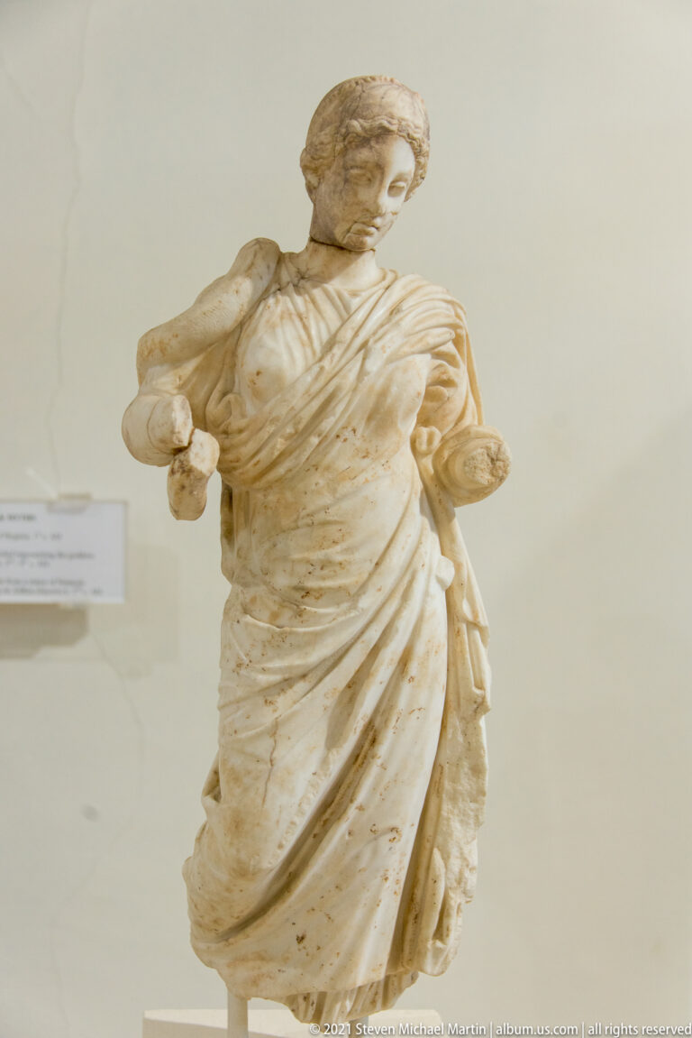 Standing marble woman in flowing drapery, with preserved head and small attributes held in both partly surviving hands.