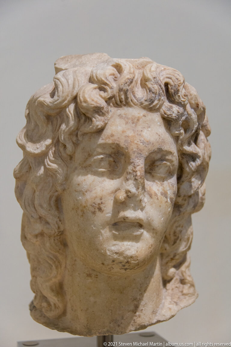 Close-up of a beardless marble youth with abundant curly hair, smooth cheeks, parted lips, and a broken neck.