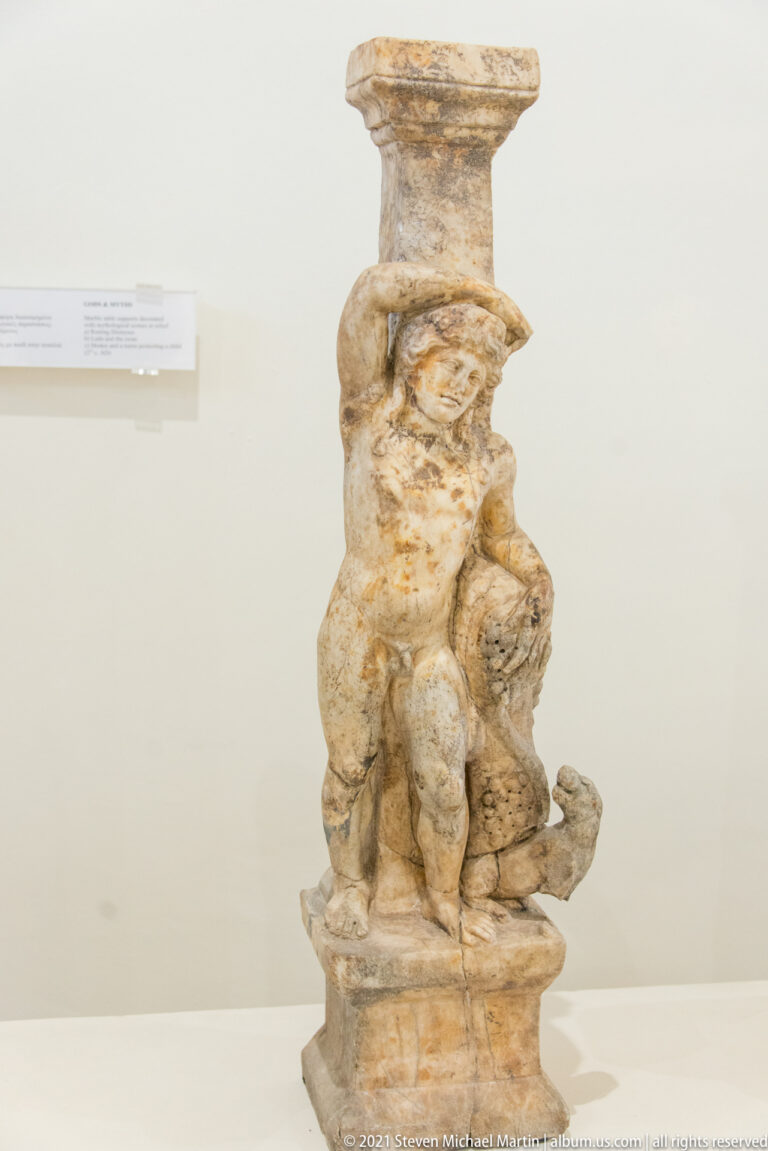 Tall carved stone support on a square base, decorated with small crouching figures and dense curling ornament beneath a molded top.