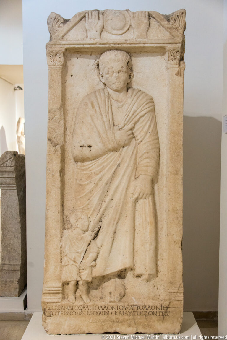 Rectangular stone stele with a standing draped male in relief and a line of Greek inscription along the bottom.