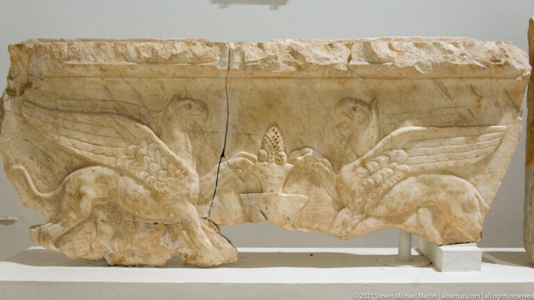 Stone relief with two winged quadrupeds facing a tall central vegetal motif on a broken rectangular slab.
