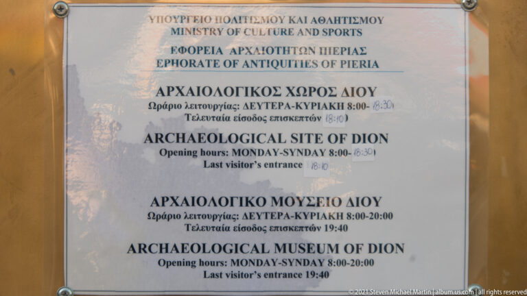 Glazed bilingual notice listing opening hours for the Archaeological Site of Dion and Archaeological Museum of Dion.