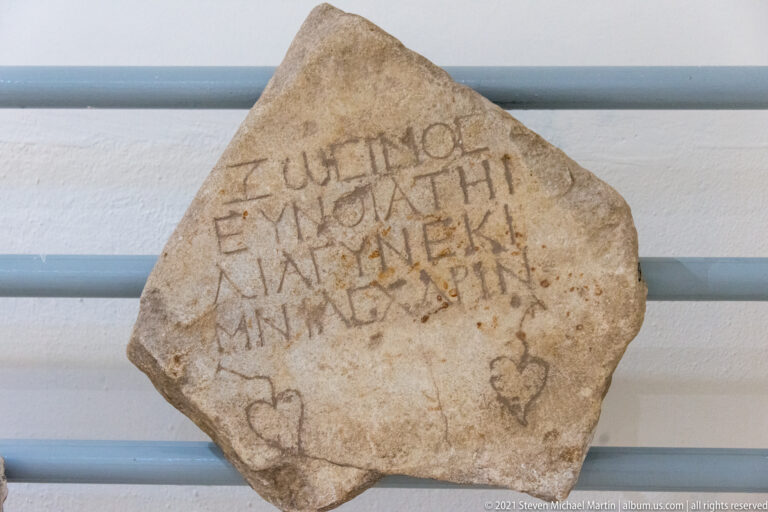 Irregular pale stone fragment bearing several lines of incised Greek letters with additional scratched marks beneath the inscription.
