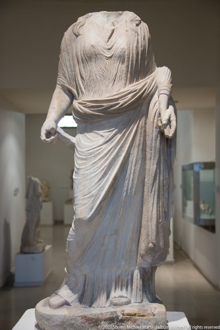 Headless marble female statue in layered, finely carved drapery, with both forearms incomplete.