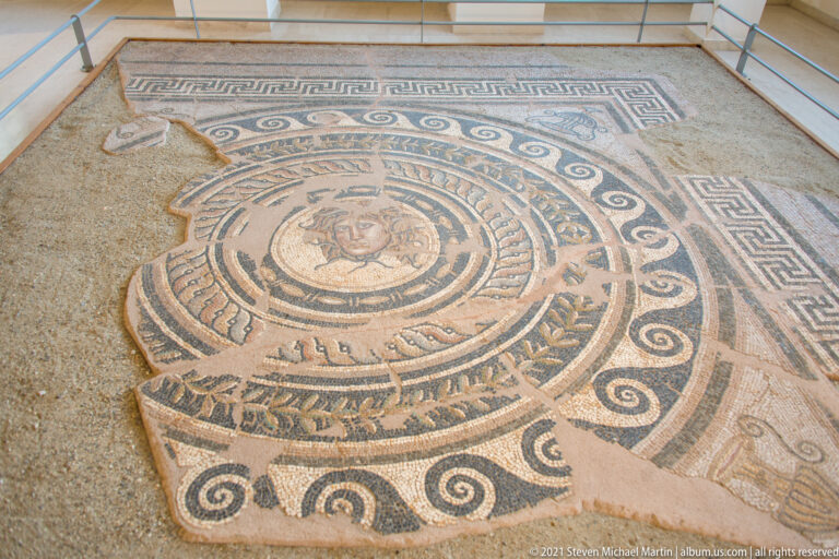 Large floor mosaic with a central female face surrounded by concentric wave, scroll, geometric, and vegetal borders.