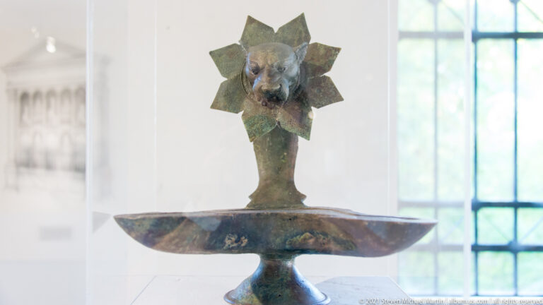 Patinated bronze pedestal vessel with a shallow bowl and tall stem ending in an animal head within a star-shaped collar.