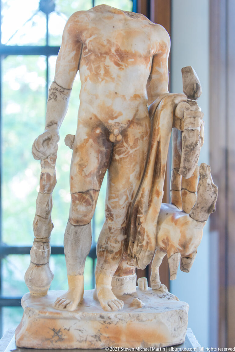 Fragmentary nude marble male standing with smaller attendant figures, a vertical support, and draped elements.