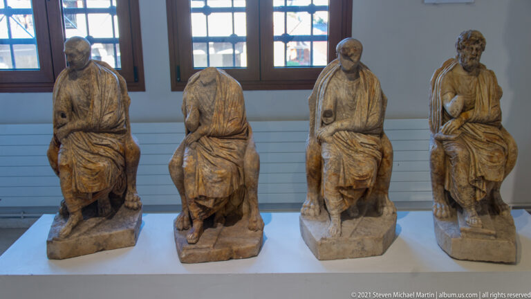 Four small seated draped male statues in a row, including bearded and headless examples with varying losses.