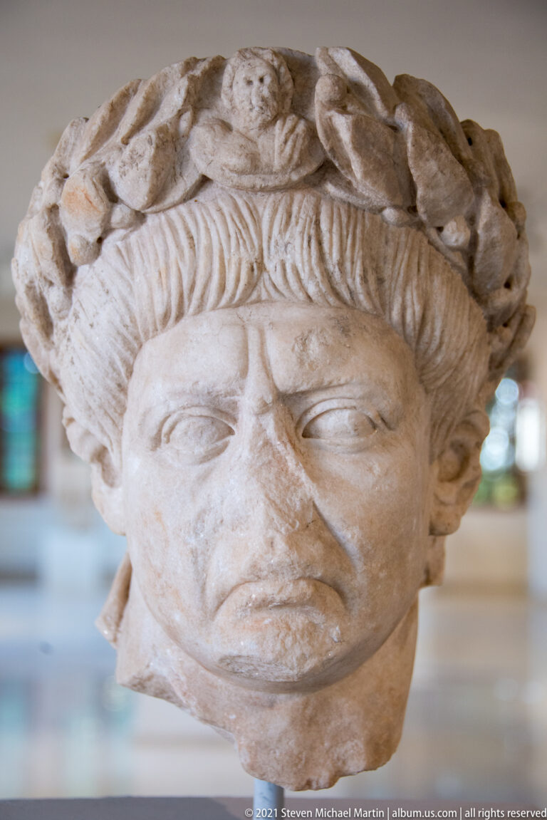 Marble female head with wavy hair and an elaborate wreath of carved leaves, flowers, and fruit-like forms.