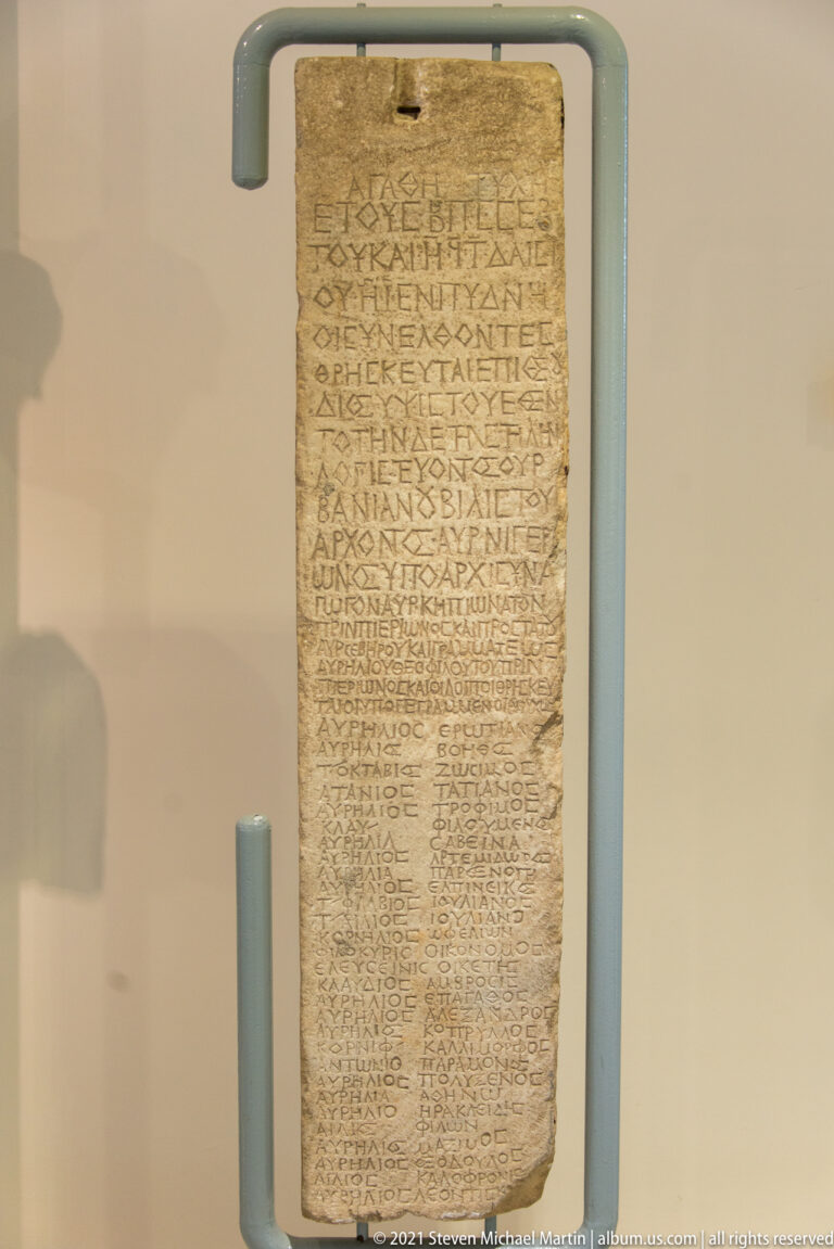 Tall rectangular stone slab covered with many lines of closely spaced Greek inscription in a museum display.