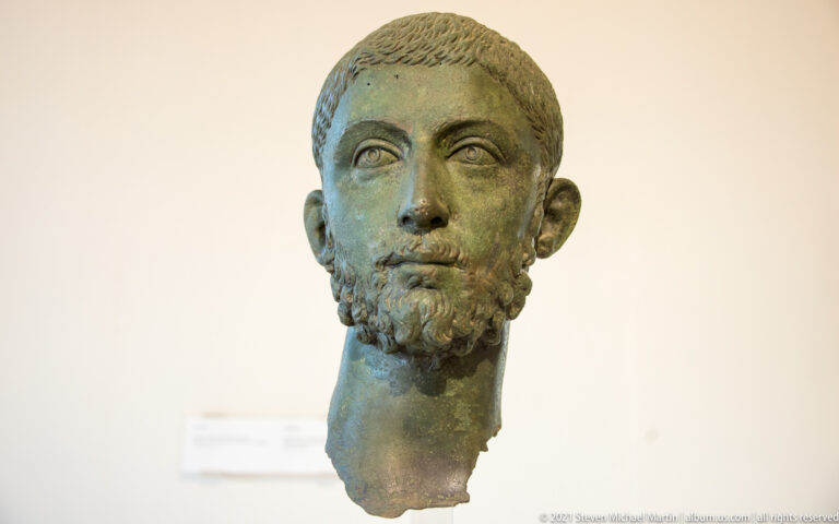 Bronze male portrait head with short hair, trimmed beard, large eyes, and an irregularly broken neck.