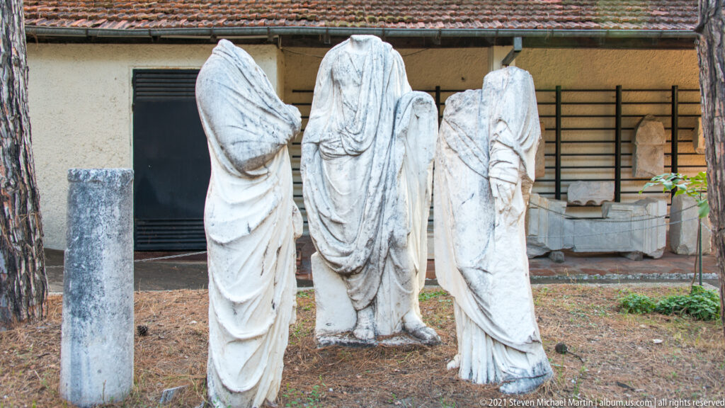 Group of tall headless draped marble statues displayed outdoors beside a museum building and trees.