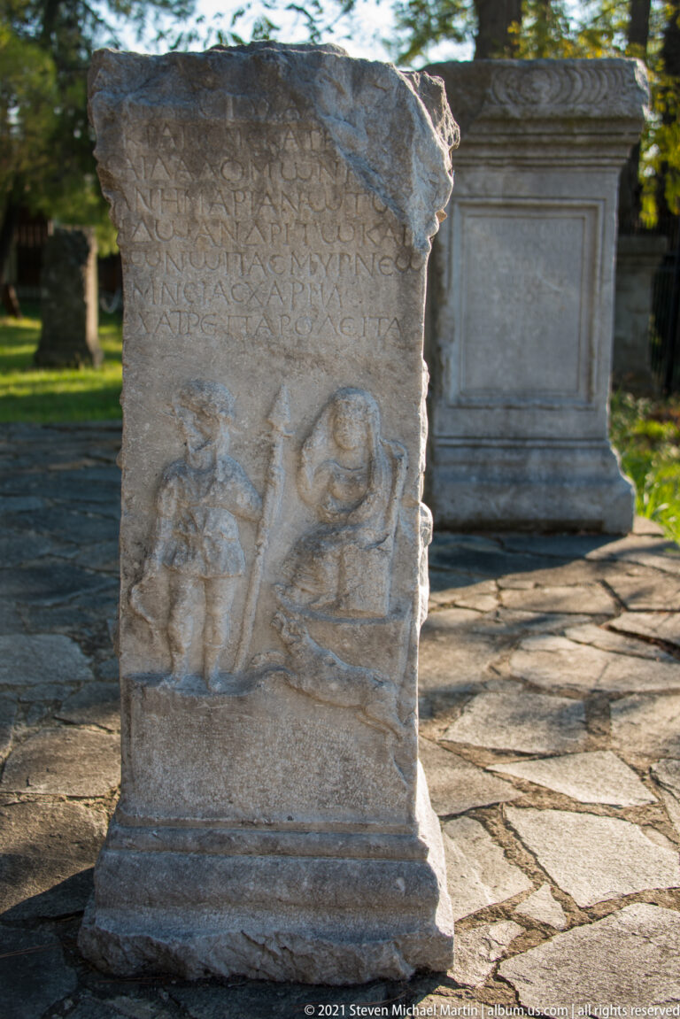 Weathered upright stone stele with Greek inscription above a recessed relief containing two facing human figures.