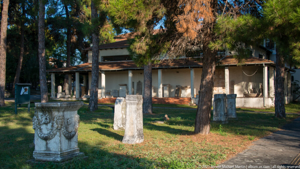 Tree-shaded museum courtyard with carved stone pedestals, upright monuments, and a low building housing additional displays.