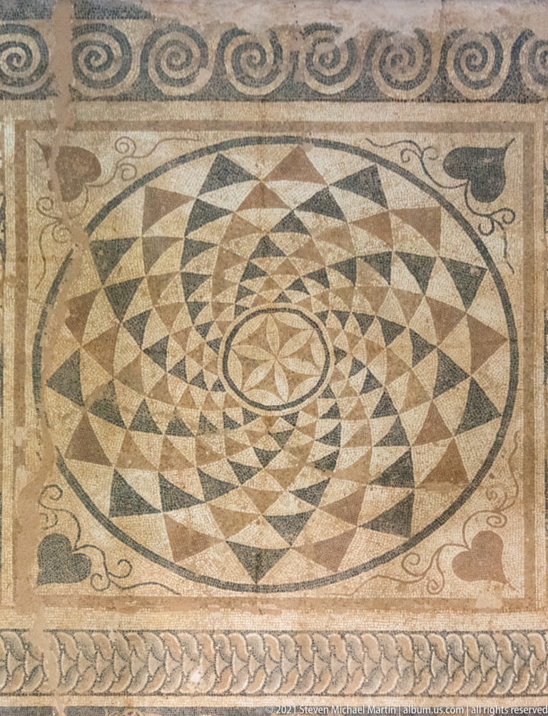 Geometric mosaic with a large circular triangular starburst, corner motifs, scrolls, and braided borders.
