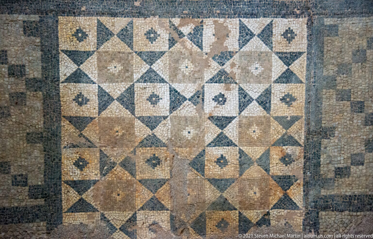 Rectangular mosaic arranged in a square grid of diamonds, triangles, and small rosettes with several damaged patches.