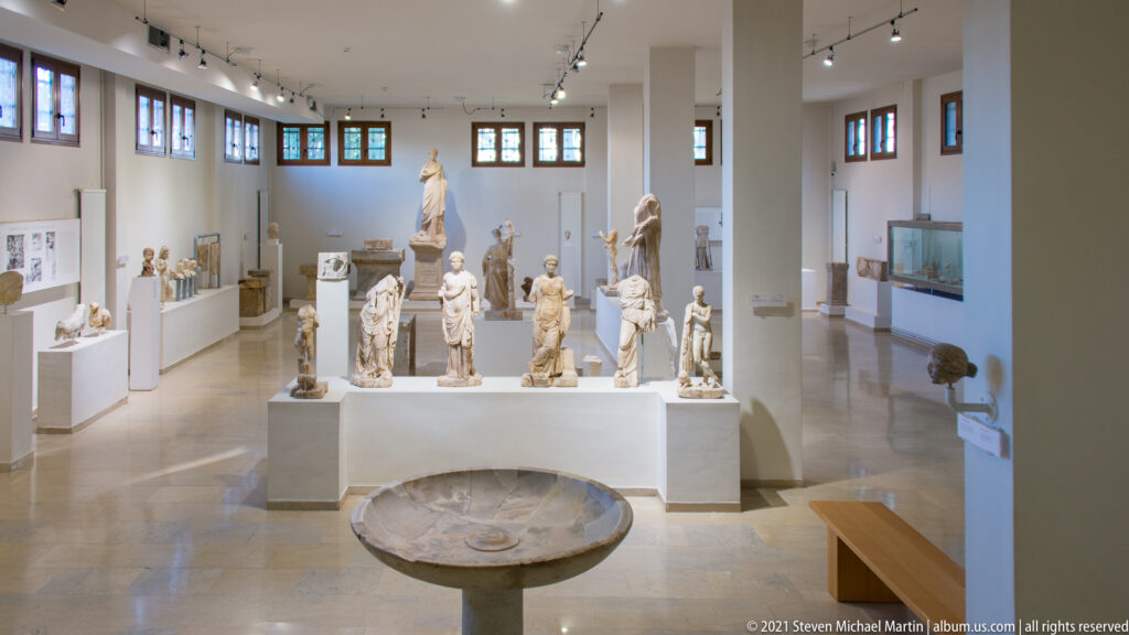 Bright museum gallery filled with marble sculptures on white pedestals, with a broad stone basin in the foreground.