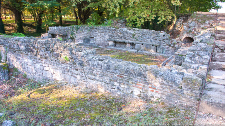 Excavated Roman bath room with low stone-and-brick walls, small wall openings, mossy floor, and an arched service passage.