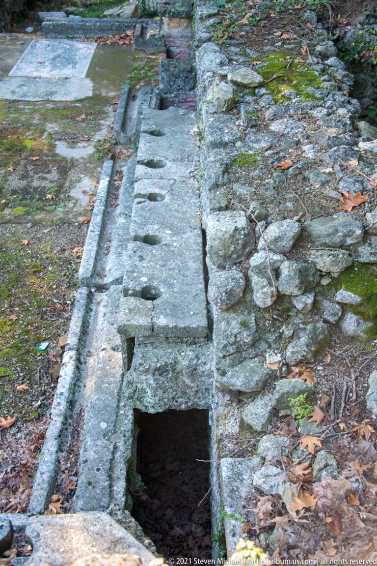 Stone-lined drainage channel with long cover slabs, circular openings, and a deep rectangular access opening.
