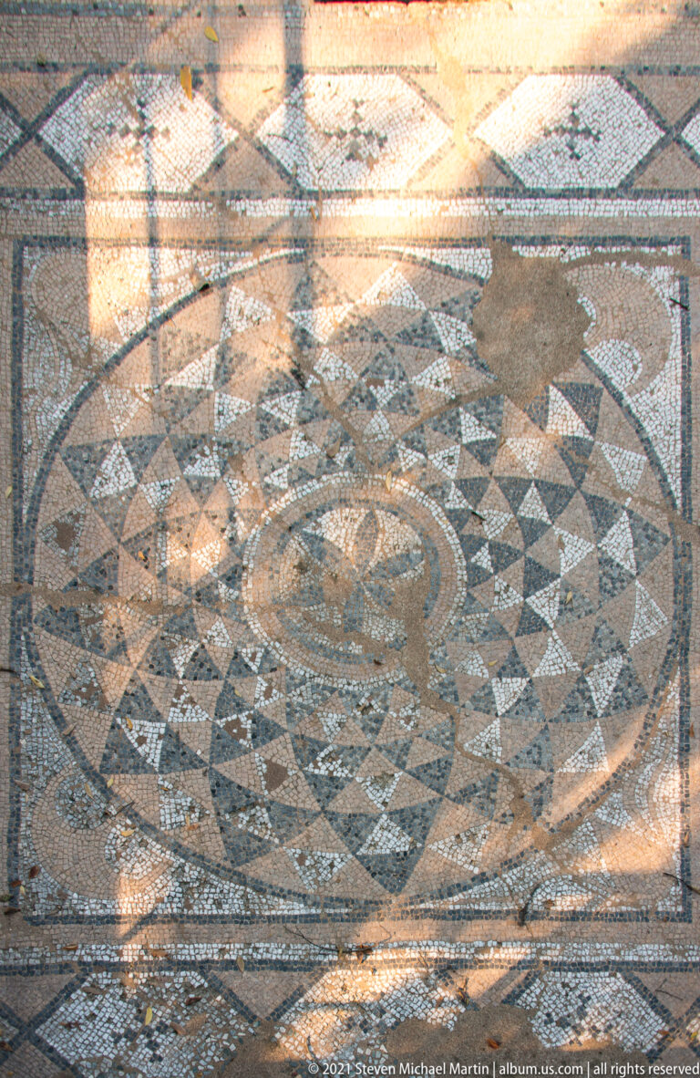 Circular geometric mosaic of interlace, triangles, and concentric bands set within a rectangular patterned pavement.