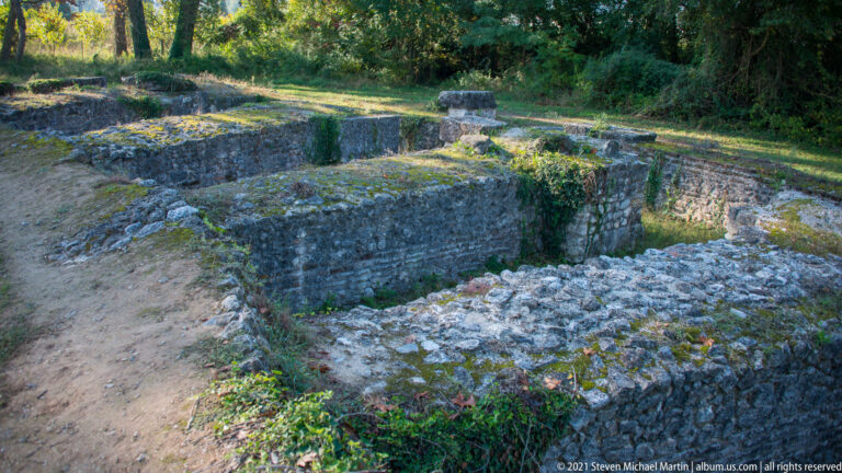 Moss-covered stone-and-brick foundations forming rectangular rooms within a wooded area of the Archaeological Park of Dion.