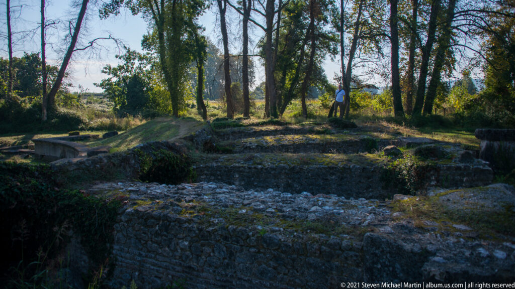 Shadowed stone foundations and excavation trenches beneath tall trees, with one visitor standing in the background.