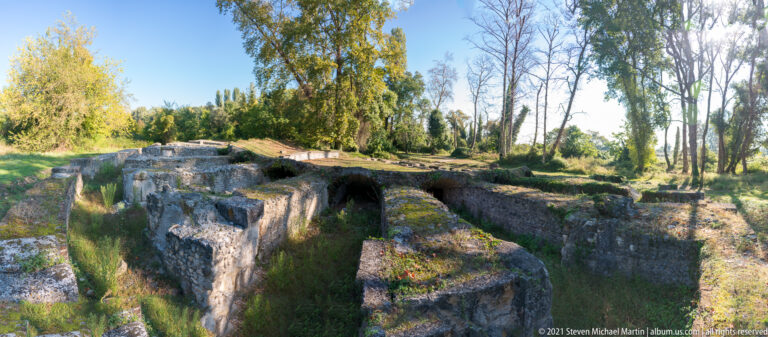 Wide view of long stone walls, corridor-like passages, and deep rectangular compartments among trees at Dion.