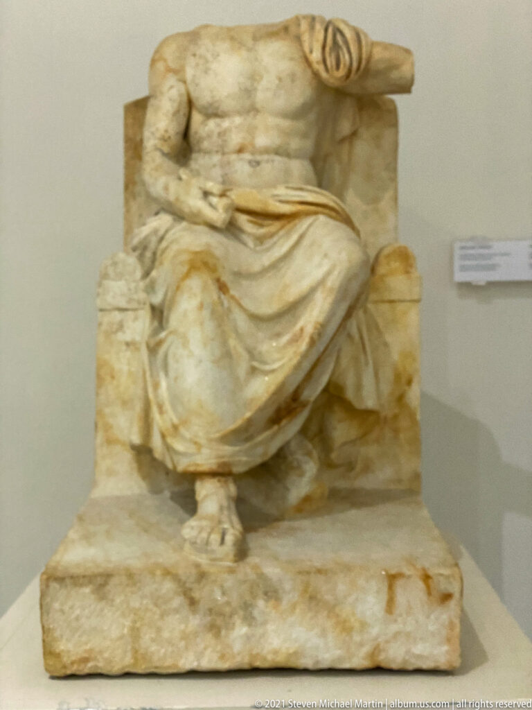 Fragmentary marble figure seated on a high-backed chair, with heavy drapery, missing head, and one preserved side element.