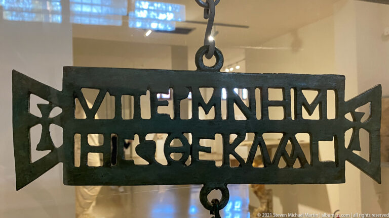 Dark patinated openwork bronze plaque with large cut-out Greek letters, rectangular frame, and triangular side ornaments.