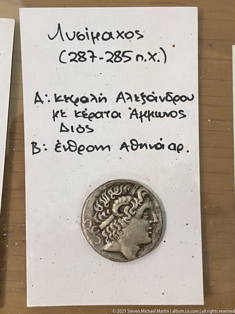 Silver coin with a right-facing male head displayed below a handwritten Greek identification note on white paper.