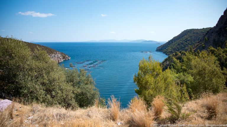 Blue coastal inlet between wooded hills, viewed across dry grasses and trees, with aquaculture pens visible offshore.