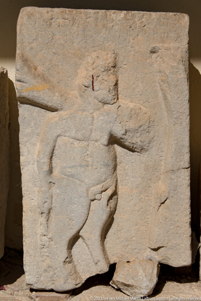 Weathered stone relief fragment with a standing profile figure extending one arm; lower portion is broken away.
