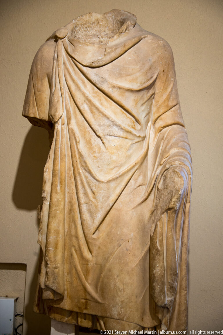 Headless marble torso wrapped in layered drapery with deep diagonal folds across the chest.