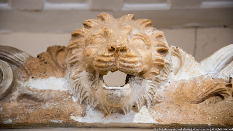 Carved stone lion head with open circular mouth and stylized mane projecting from an architectural fragment.