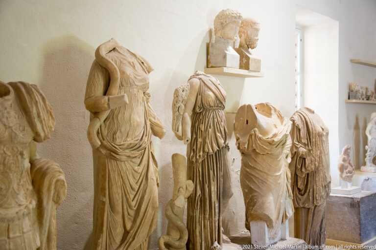 Museum gallery with several headless draped marble statues and additional sculptural fragments displayed behind them.
