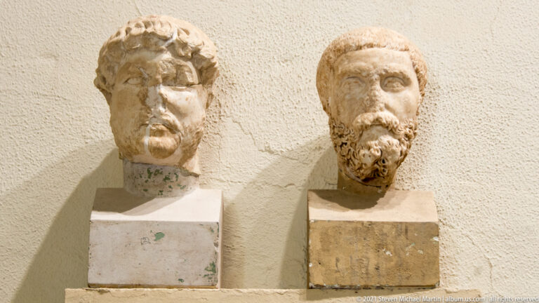 Two bearded marble male heads mounted side by side against a museum wall.
