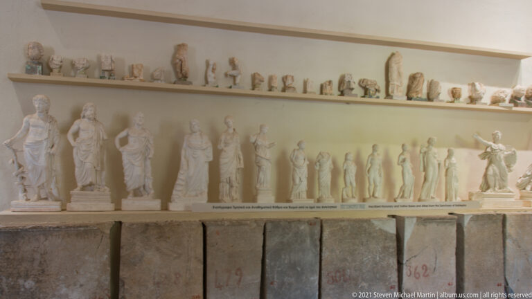 Museum shelves filled with small sculptural fragments above a row of standing marble statuettes and stone blocks.