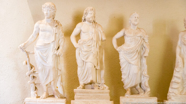Three standing marble statuettes; the headless figure at left has a serpent coiled around a staff.