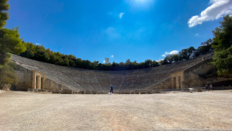 Wide view from the orchestra toward the semicircular stone seating of the Ancient Theatre of Epidaurus under blue sky.