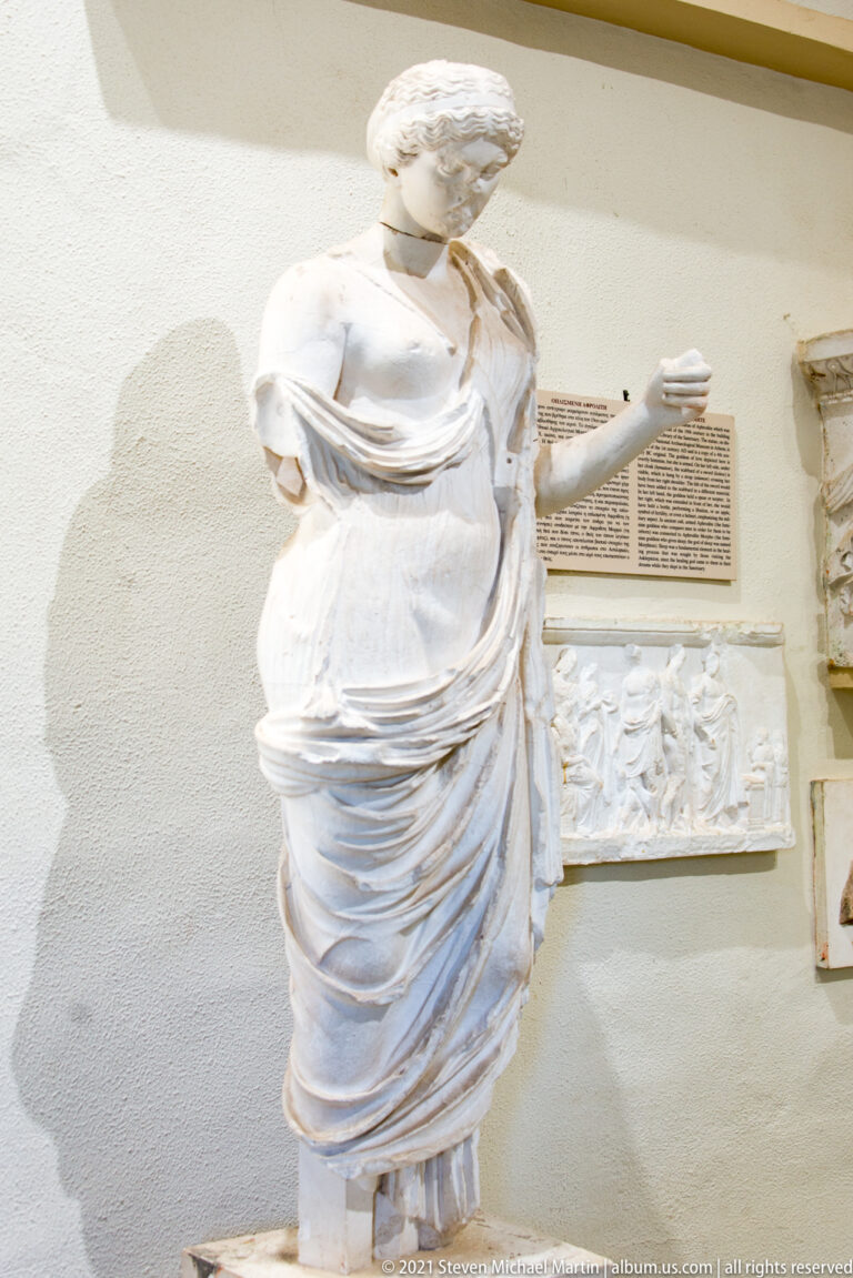 Standing female marble statue in layered drapery, with damaged head and one surviving arm extended outward.