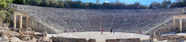 Elevated frontal view of the Ancient Theatre of Epidaurus with circular orchestra, stone seating, side passages, and visitors.