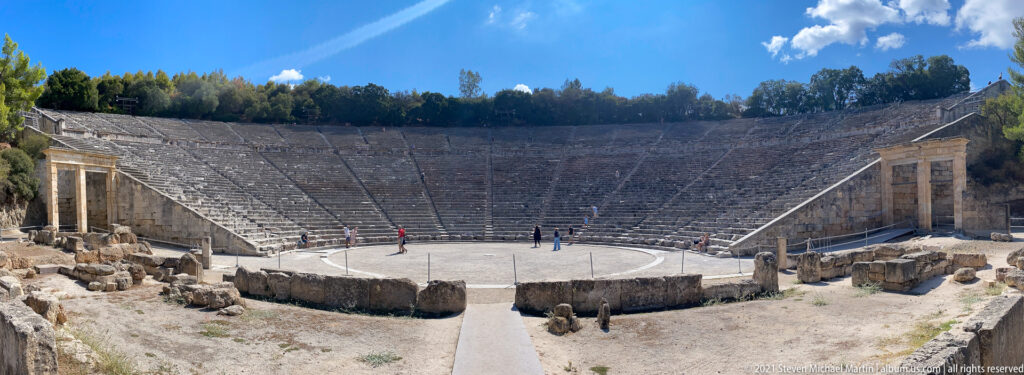 Wide elevated view of the Ancient Theatre of Epidaurus with orchestra, seating, visitors, and stone ruins in the foreground.