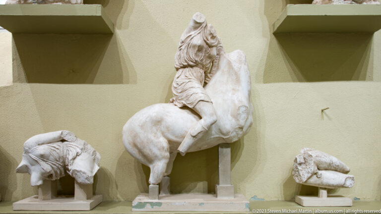 Fragmentary marble horse and rider mounted upright, with separate sculptural fragments displayed on supports at both sides.