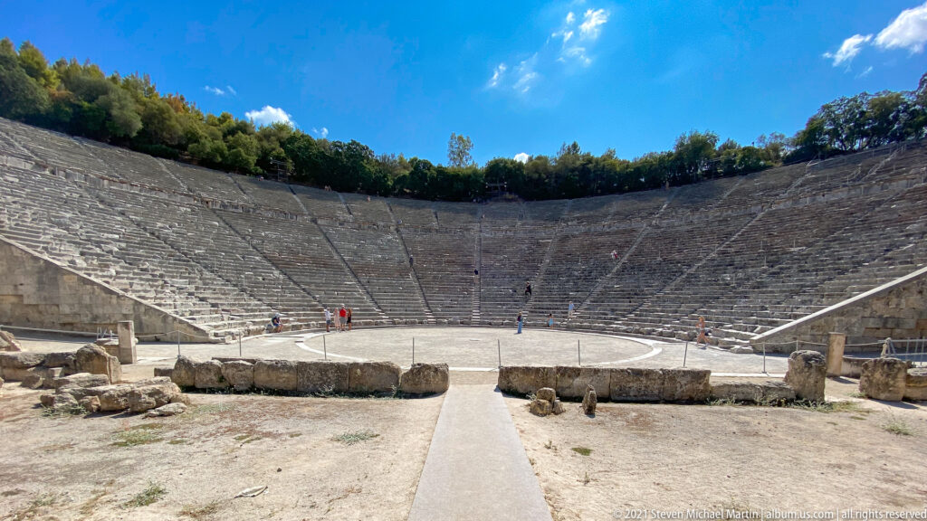 Wide stage-side view across the circular orchestra toward the steep stone seating of the ancient theatre.