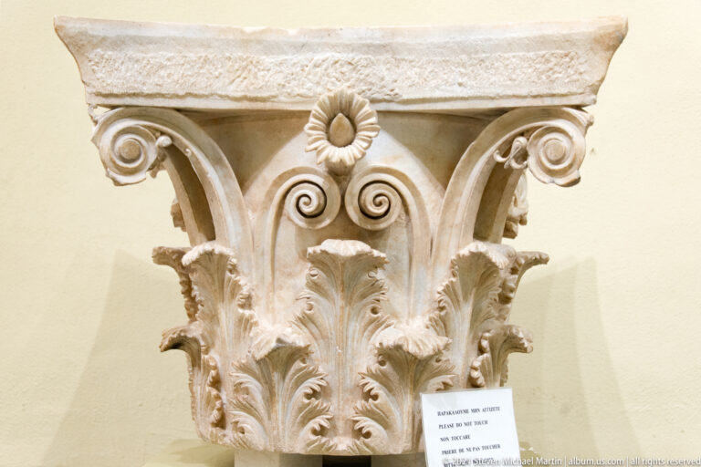 Ornate marble Corinthian capital with acanthus leaves, spiral volutes, and a central rosette in a museum display.