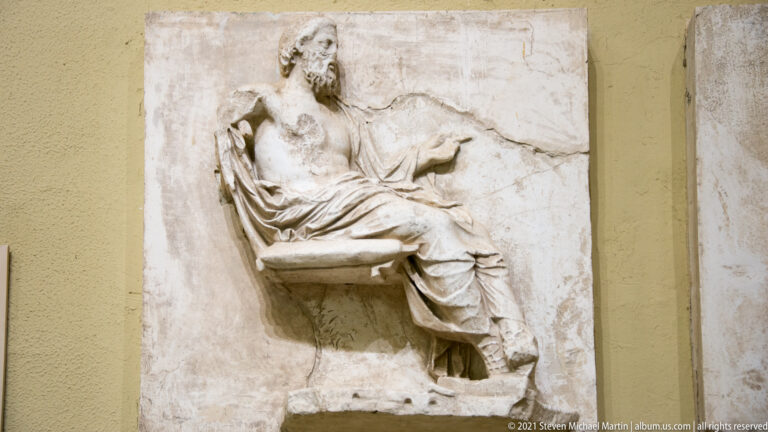 Marble relief of a seated bearded man in drapery, with one arm raised against a shallow carved background.